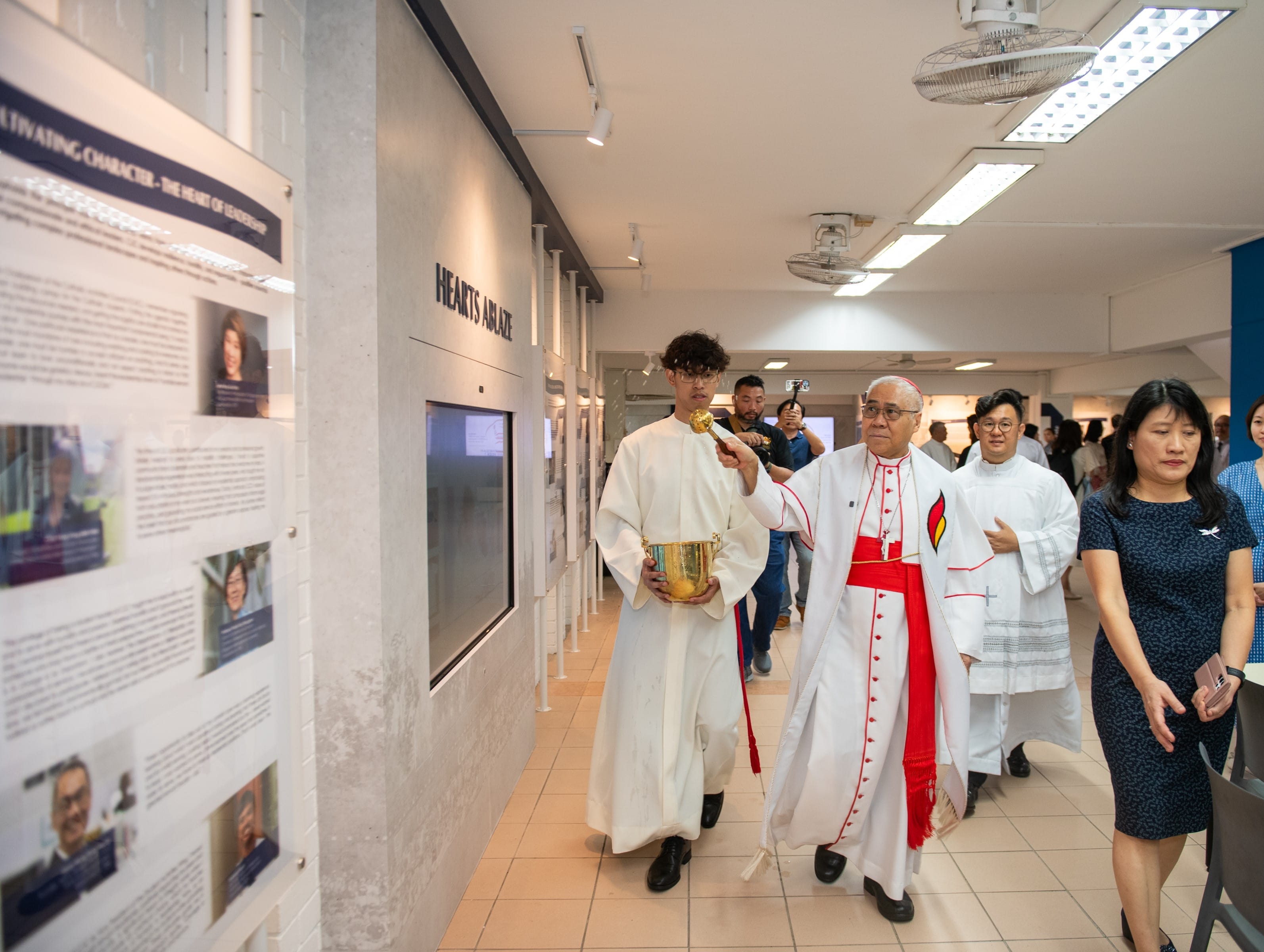 A Catholic cardinal in red and white vestments walks through the Catholic Junior College Heritage Gallery during a blessing ceremony, accompanied by an altar server in white robes carrying a golden holy water vessel and aspergillum. Other clergy and guests follow behind. The 'Hearts Ablaze' signage and heritage display panels are visible along the corridor wall.